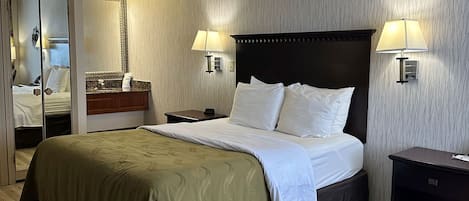 Hotellihuone, jossa on sänky, yöpöydällä olevat lamput, yöpöytä, peili ja puulattia.