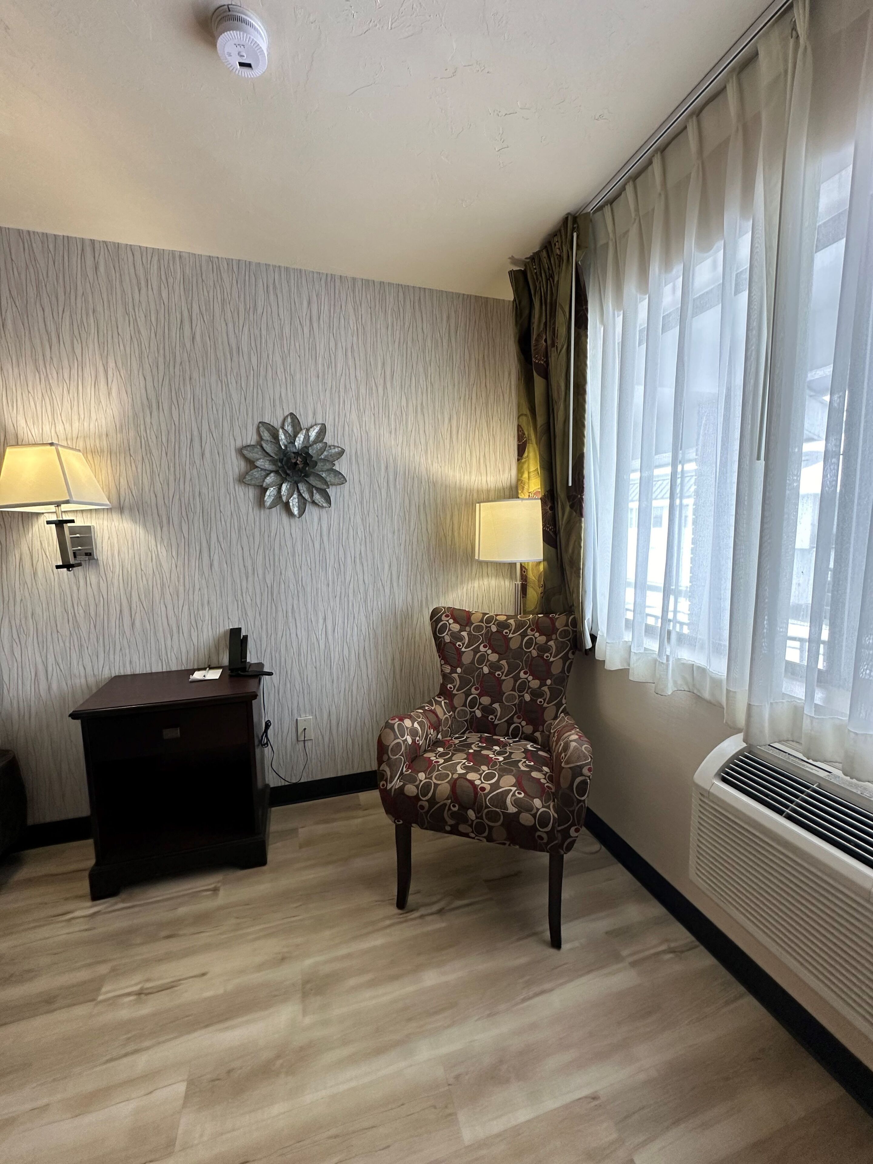 A room with a patterned chair, a desk with a lamp, a decorative wall piece, and a window with curtains.