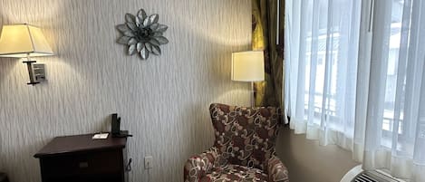 A room with a patterned chair, a desk with a lamp, a decorative wall piece, and a window with curtains.