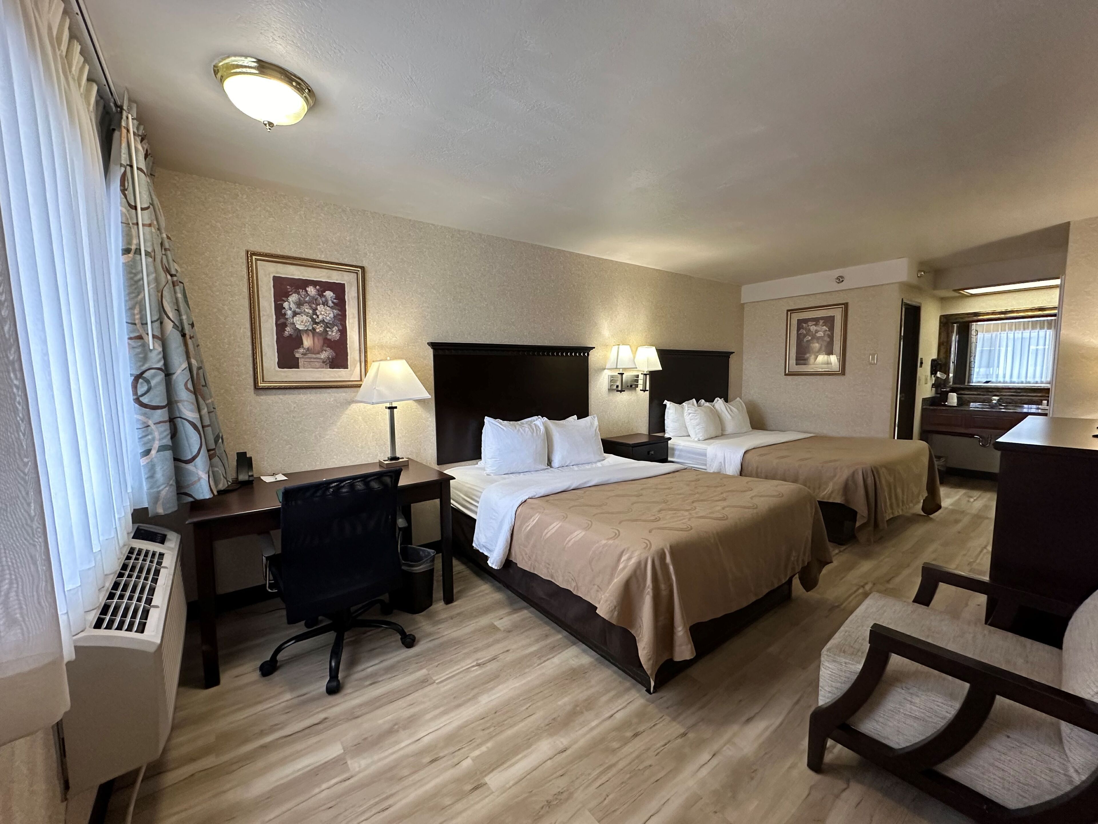 Quarto de hotel com duas camas, uma mesa de escritório, uma cadeira, uma luminária de mesa, uma mesa de cabeceira e uma janela com cortinas.