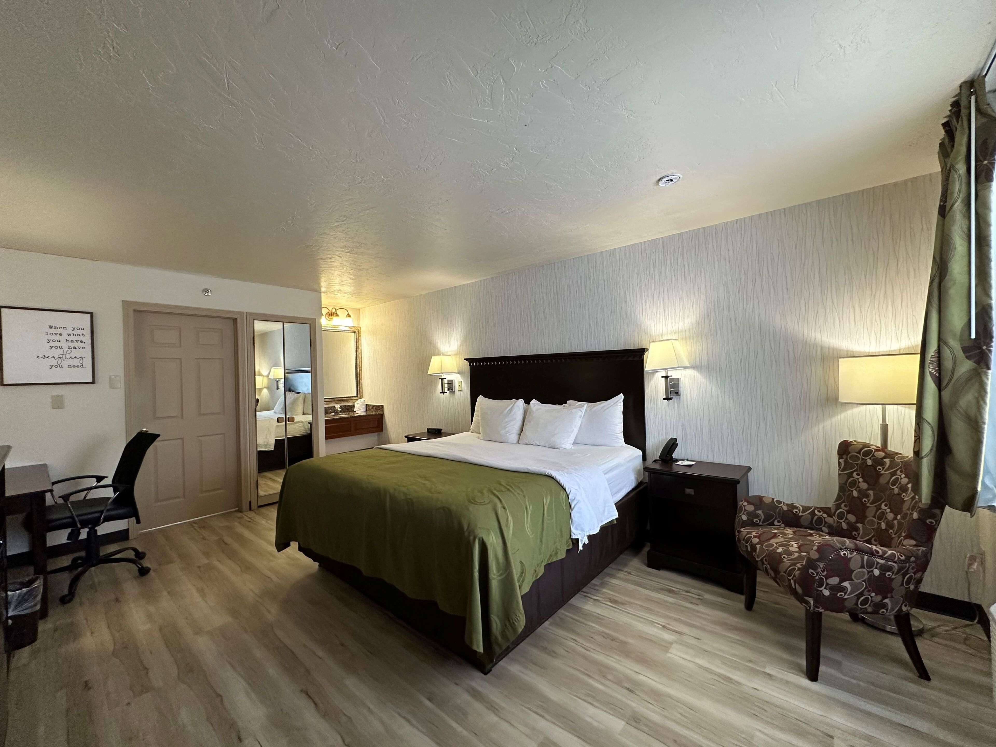 Una habitación de hotel con cama, mesitas de noche, una silla, escritorio, espejo y una lámpara en la pared.