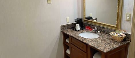 A bathroom with a granite countertop, a white sink, a large mirror with a gold frame, a coffee maker, and a basket of toiletries.