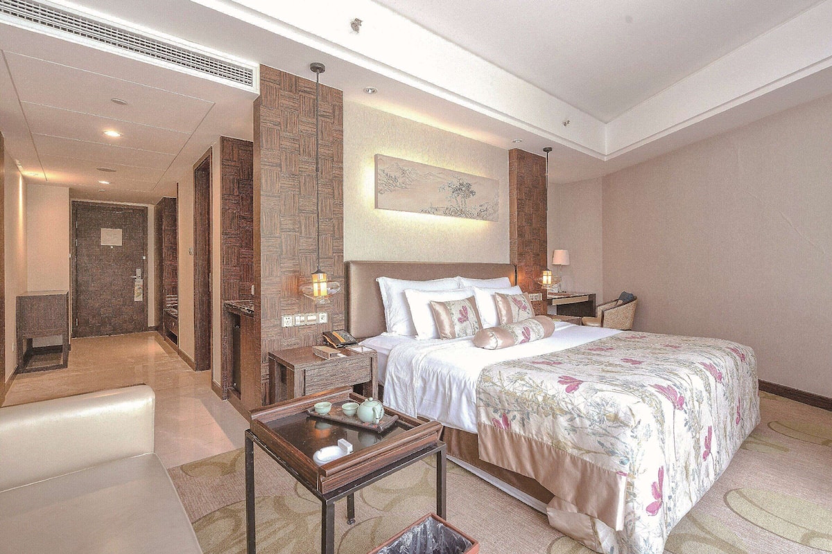 Une chambre d’hôtel avec un grand lit, des tables de chevet, une petite table, une œuvre d’art accrochée au mur et un couloir menant à une autre pièce.