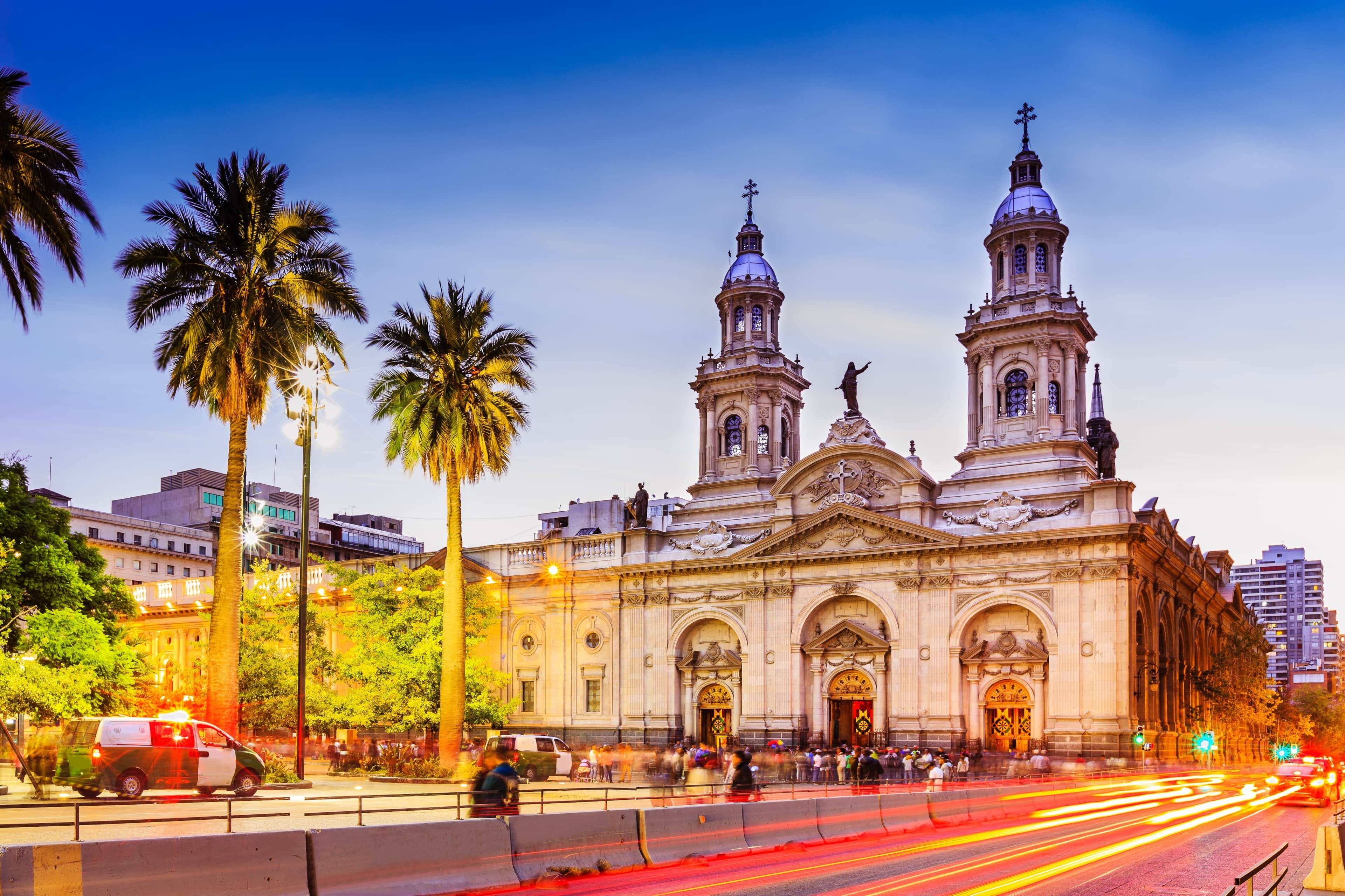 A historic building with two prominent towers and a central dome, surrounded by palm trees and a busy street with light trails.