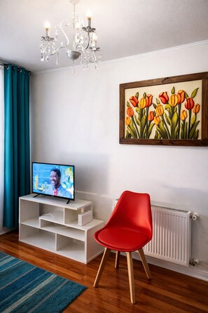 A modern living room with a television, a red chair, a chandelier, and a tulip painting on the wall.