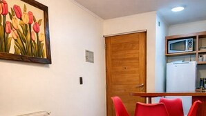 A room with a wooden door, a dining table with red chairs, a microwave, a refrigerator, and a painting of tulips on the wall.