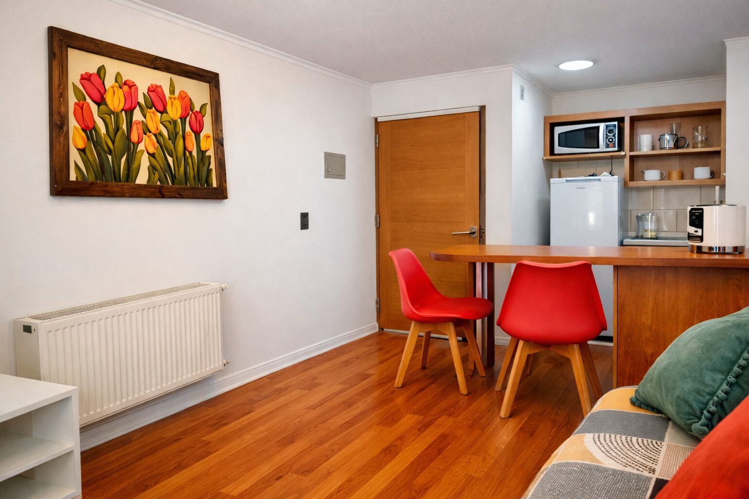 Une cuisine compacte équipée d’un four à micro-ondes, d’un grille-pain et d’étagères où sont rangés les ustensiles de cuisine. On y trouve une porte en bois, un radiateur et un tableau représentant des tulipes accroché au mur. Deux chaises rouges sont placées autour d’une table en bois.
