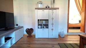 Un salon équipé d’un téléviseur, doté d’un plancher en bois, comprenant un meuble où sont exposés des objets décoratifs, et offrant une vue sur la chambre à coucher.