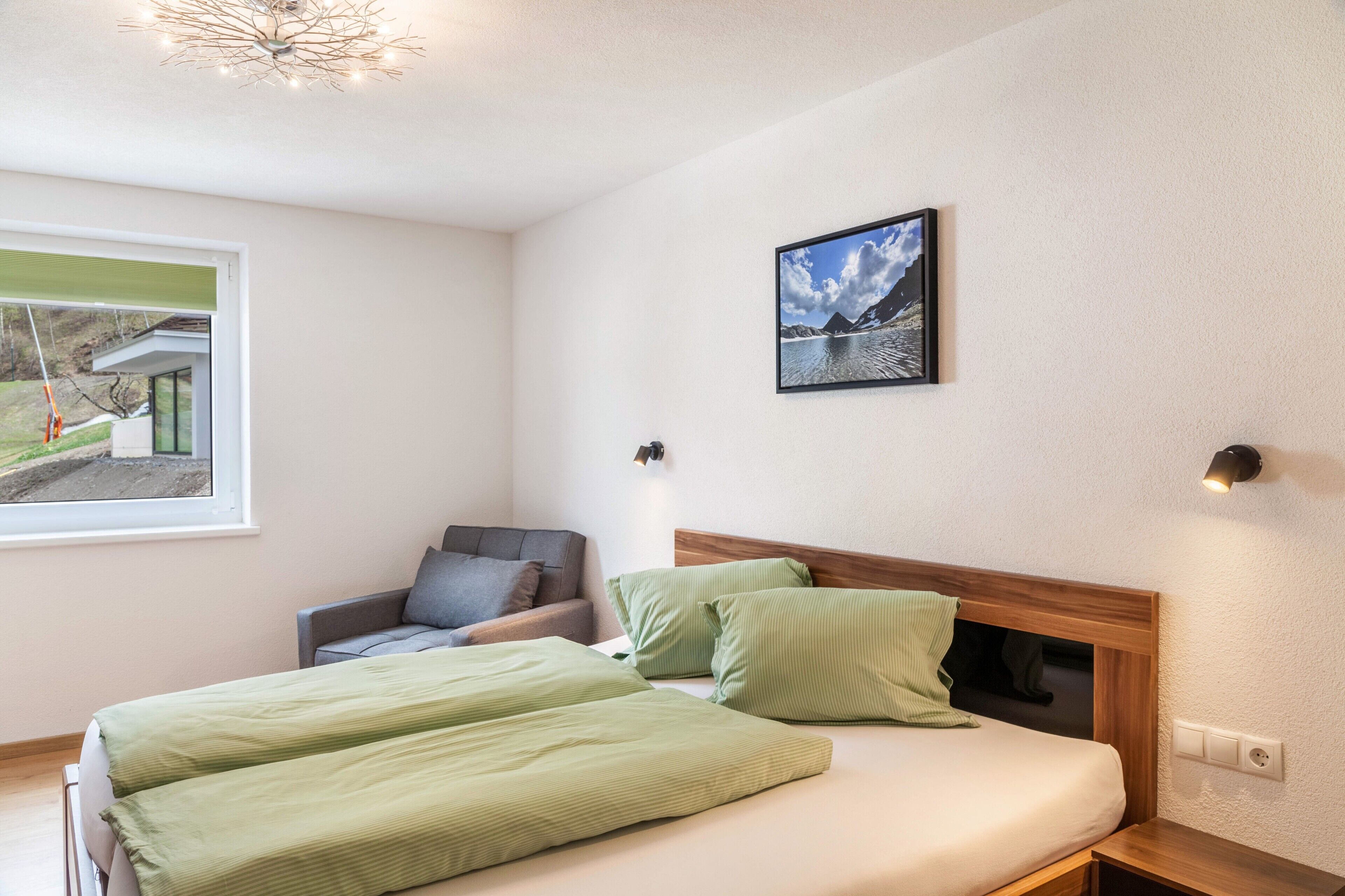Ein Schlafzimmer mit Bett, einem Sessel, einem Wandbild und einem Fenster mit Jalousien.