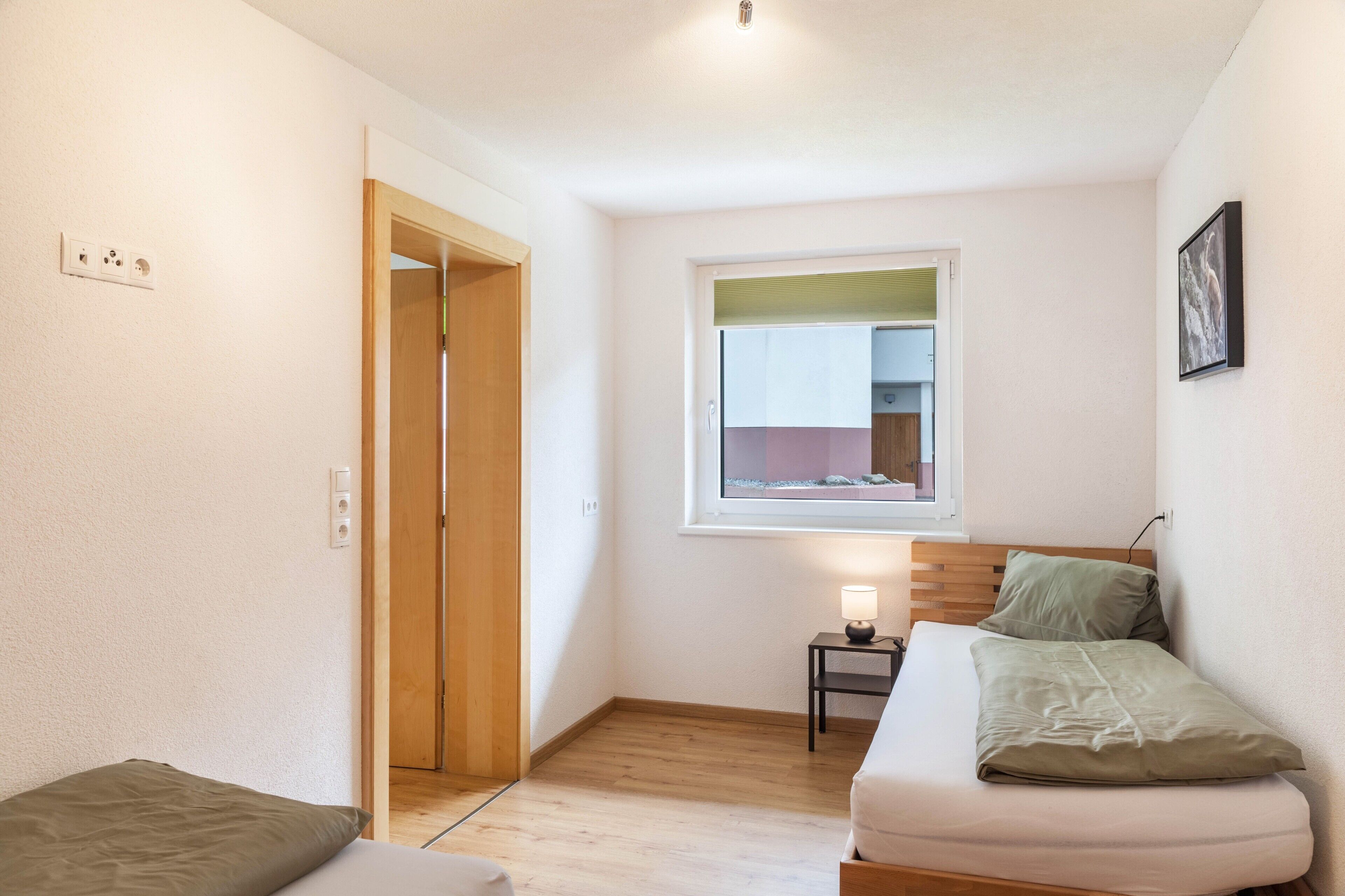 Ein kleiner, gut beleuchteter Raum mit einem Bett, einem Nachttisch mit Lampe, einem Fenster mit Ausblick und einer Tür zu einem anderen Raum.
