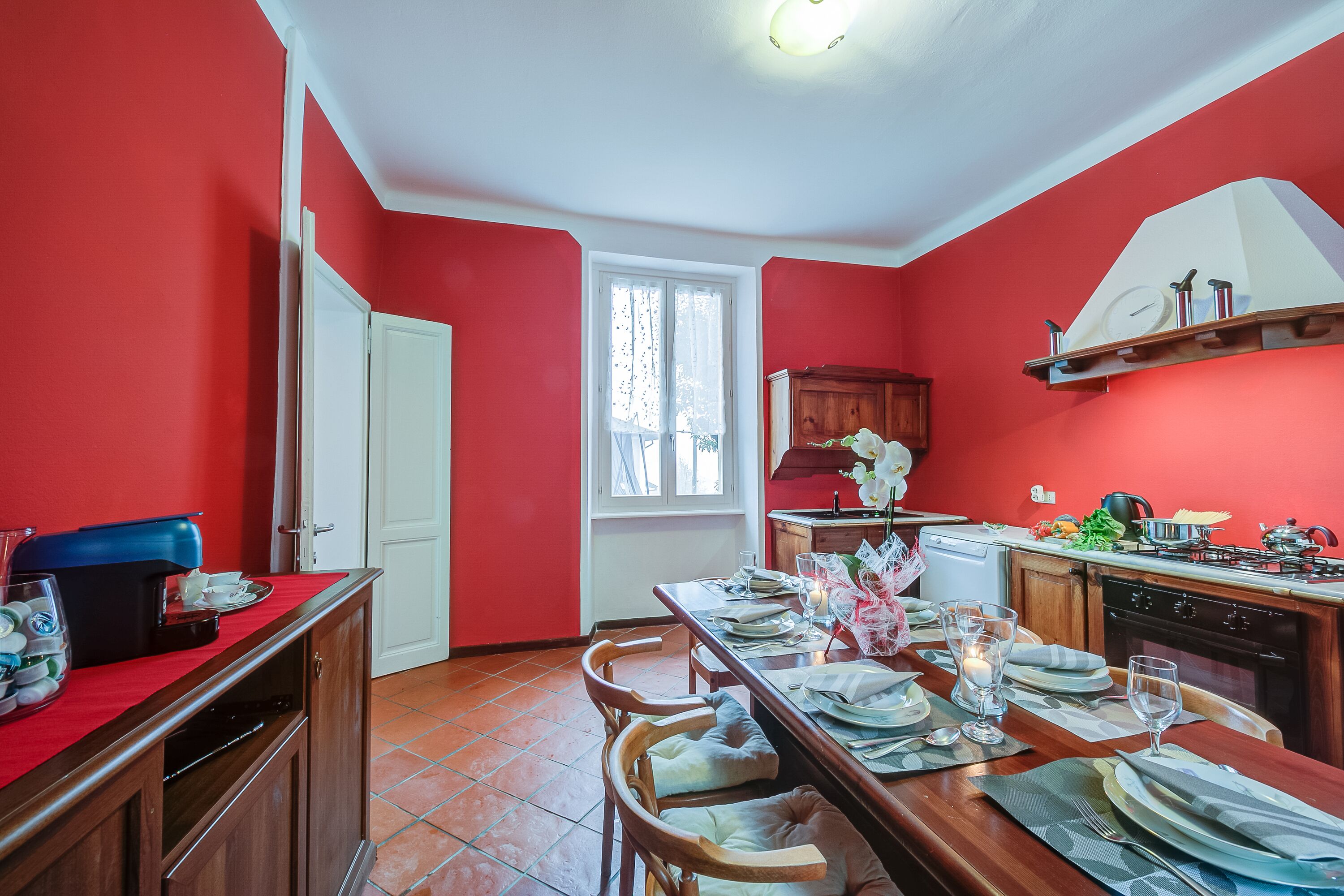 A red-walled kitchen with a dining area set for four, including a coffee machine, a sink, and a stove.