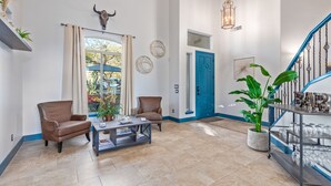 Amplia sala de estar con un sofá de cuero marrón, una mesita de centro y una planta grande en maceta. Se ve una puerta azul y una escalera con barandilla al fondo.