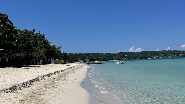 A beach with clear water, a sandy shore, and a distant view of a town. - ARA OASIS - loft condo, 5 mins walk to beach (Negril)