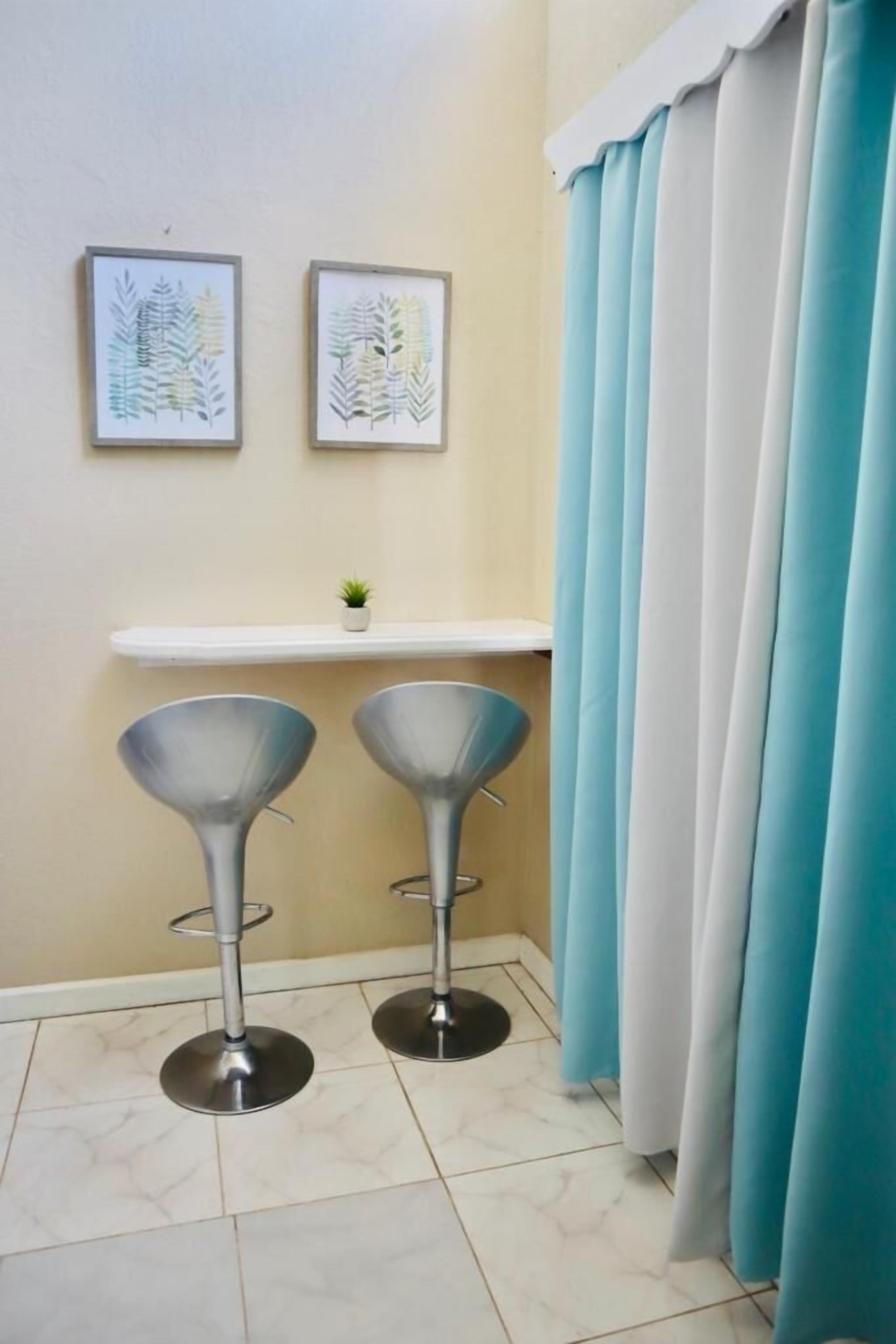 A corner of a room with two bar stools, a small shelf with a plant, and a pair of curtains.