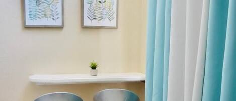 A corner of a room with two bar stools, a small shelf with a plant, and a pair of curtains.