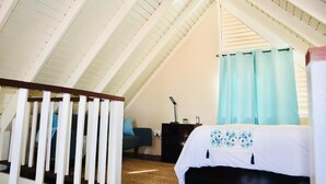 A bedroom with a sloped ceiling, a bed with a floral bedspread, a nightstand, a lamp, and a window with curtains. - ARA OASIS - loft condo, 5 mins walk to beach (Negril)