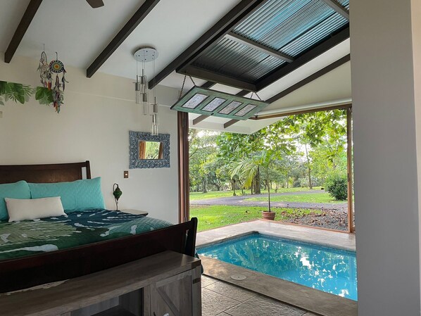 A bedroom with a bed, a nightstand, a window with a view of a swimming pool and garden.