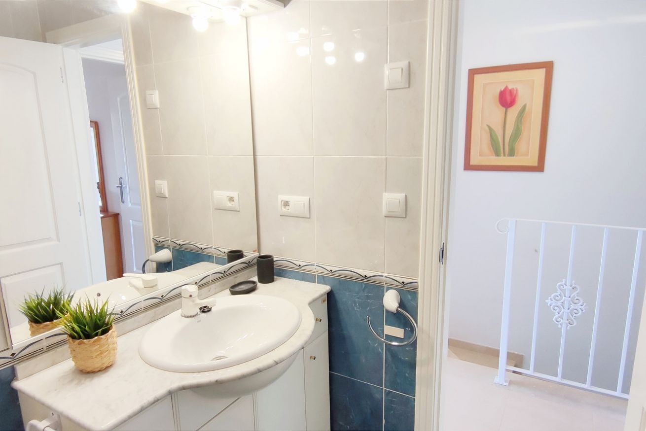 Un baño con un lavamanos blanco, un espejo, una planta en maceta, un dispensador de papel higiénico y un cuadro en la pared.