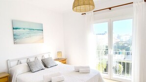 A bedroom with a bed, pillows, a bedside table, a lamp, and a view of the outside.