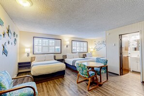 Two single beds with white linens and blue accents, a kitchenette, and a window with blinds.