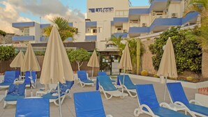 A poolside area with blue lounge chairs and beige umbrellas, a building with a sign reading "Lutesa", and a landscaped garden with palm trees. - Lufesa Apartments (Mogan)