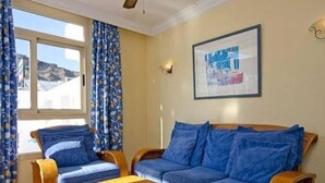 A room with a sofa and two armchairs, a wooden table, a framed picture on the wall, and a window with curtains. - Lufesa Apartments (Mogan)