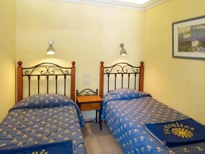 Una habitación con dos camas individuales, cada una con cabecera y mesita de noche. Dos lámparas de pared iluminan las camas.