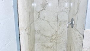 A shower with a white tiled wall and floor, a showerhead, and a drain. - The Doze Off (Houston)