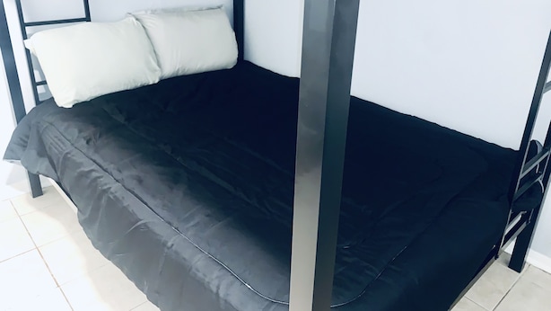 A single bed with a black mattress and white pillows in a room with white walls and a window with blinds. - The Doze Off (Houston)
