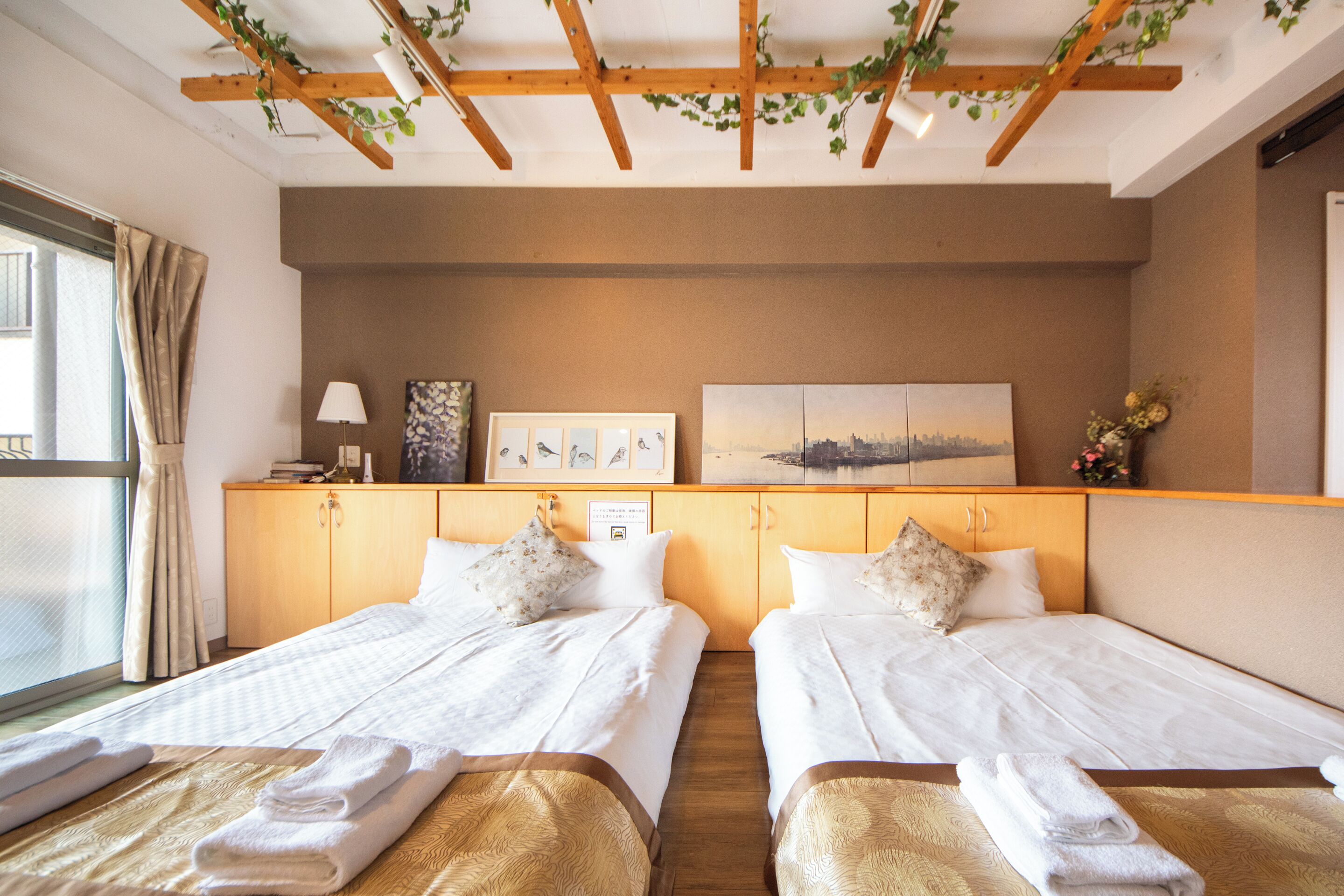 A hotel room with two beds, a wooden ceiling, a window with curtains, and artwork on the wall.