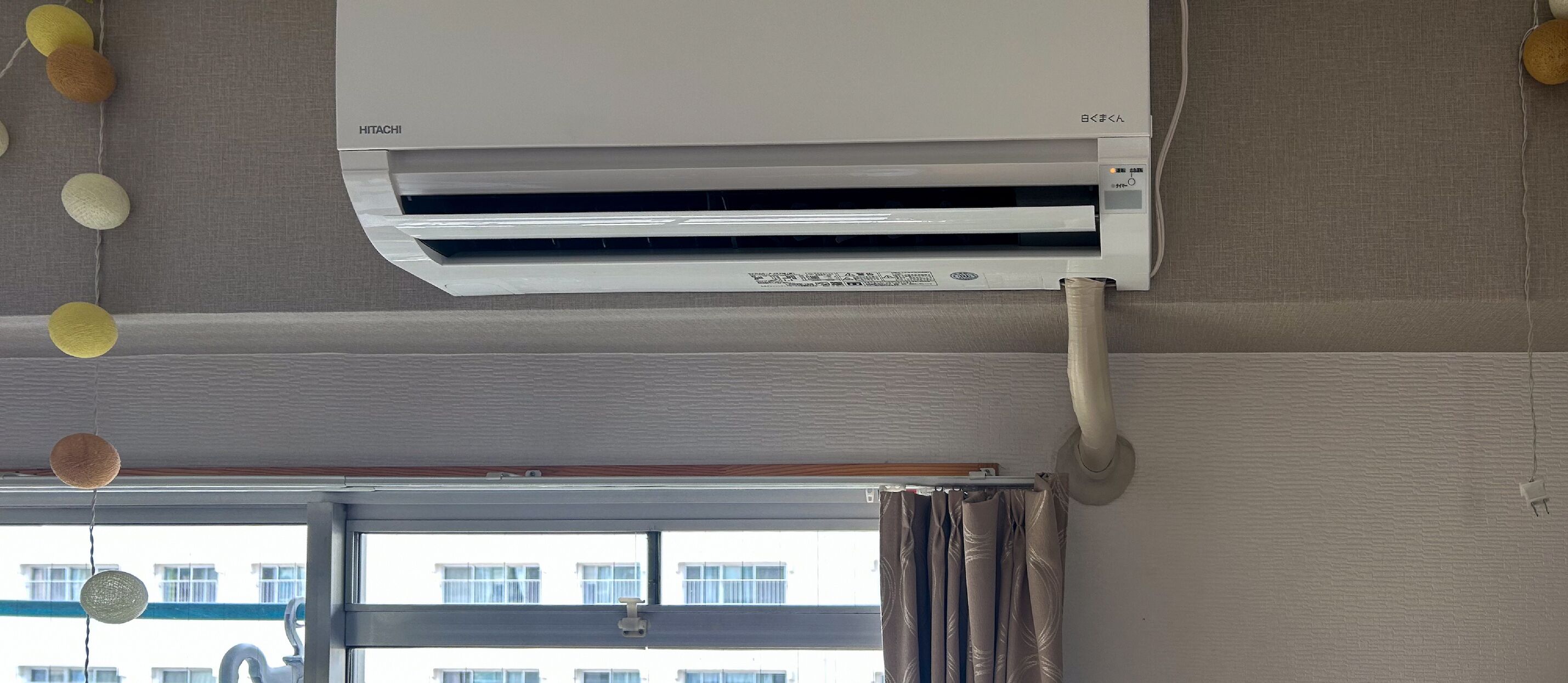Una habitación con aire acondicionado instalado en el techo, una ventana con cortinas y vistas a edificios.