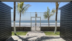Vista desde un porche techado hacia una playa, con un muro de piedra y palmeras.