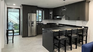 Una cocina moderna con armarios oscuros, un refrigerador de acero inoxidable, una isla grande con taburetes de bar y vistas a través de una puerta de vidrio.
