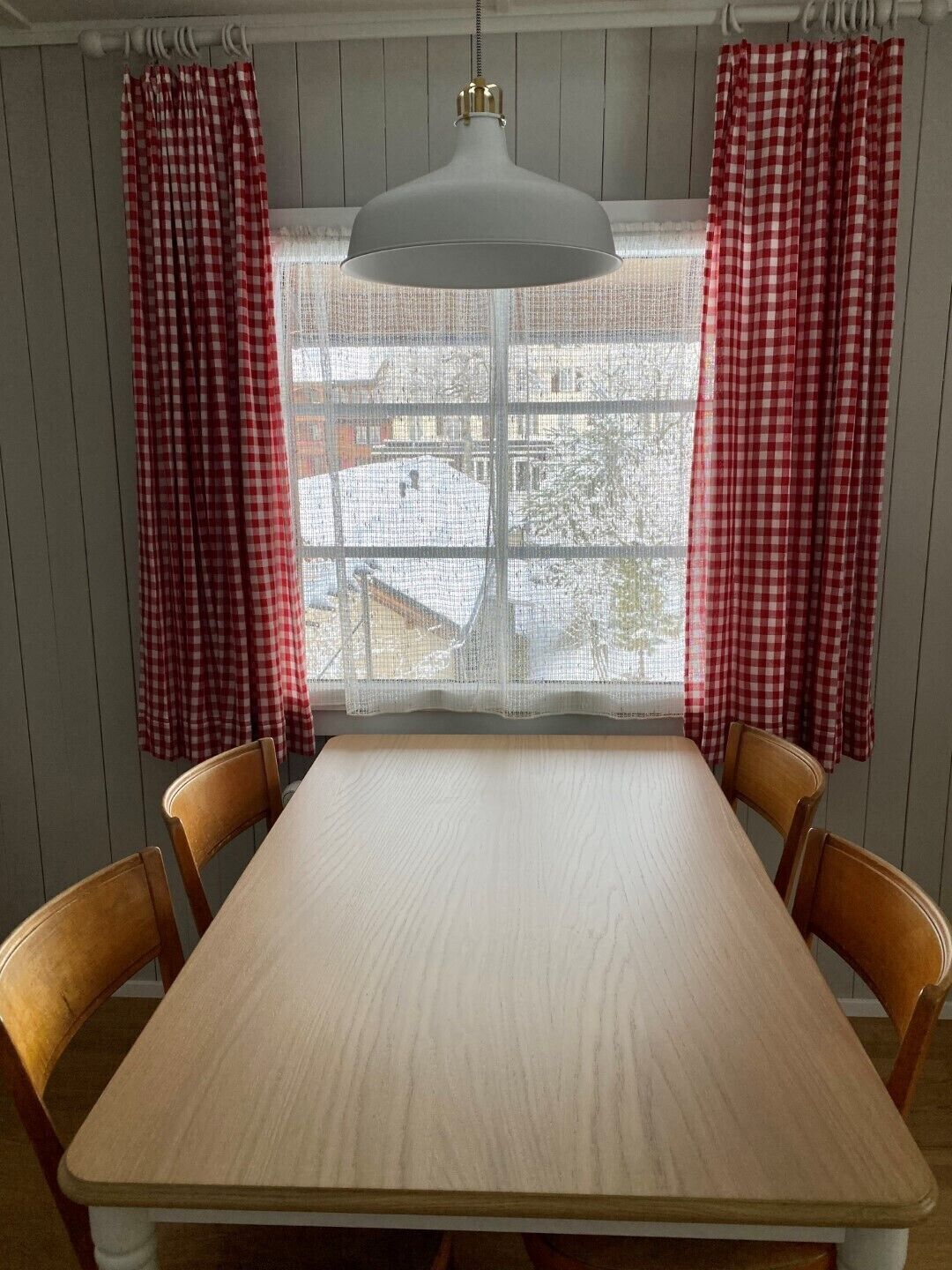 Comedor con mesa de madera, dos sillas, lámpara colgante y cortinas a cuadros rojos y blancos.