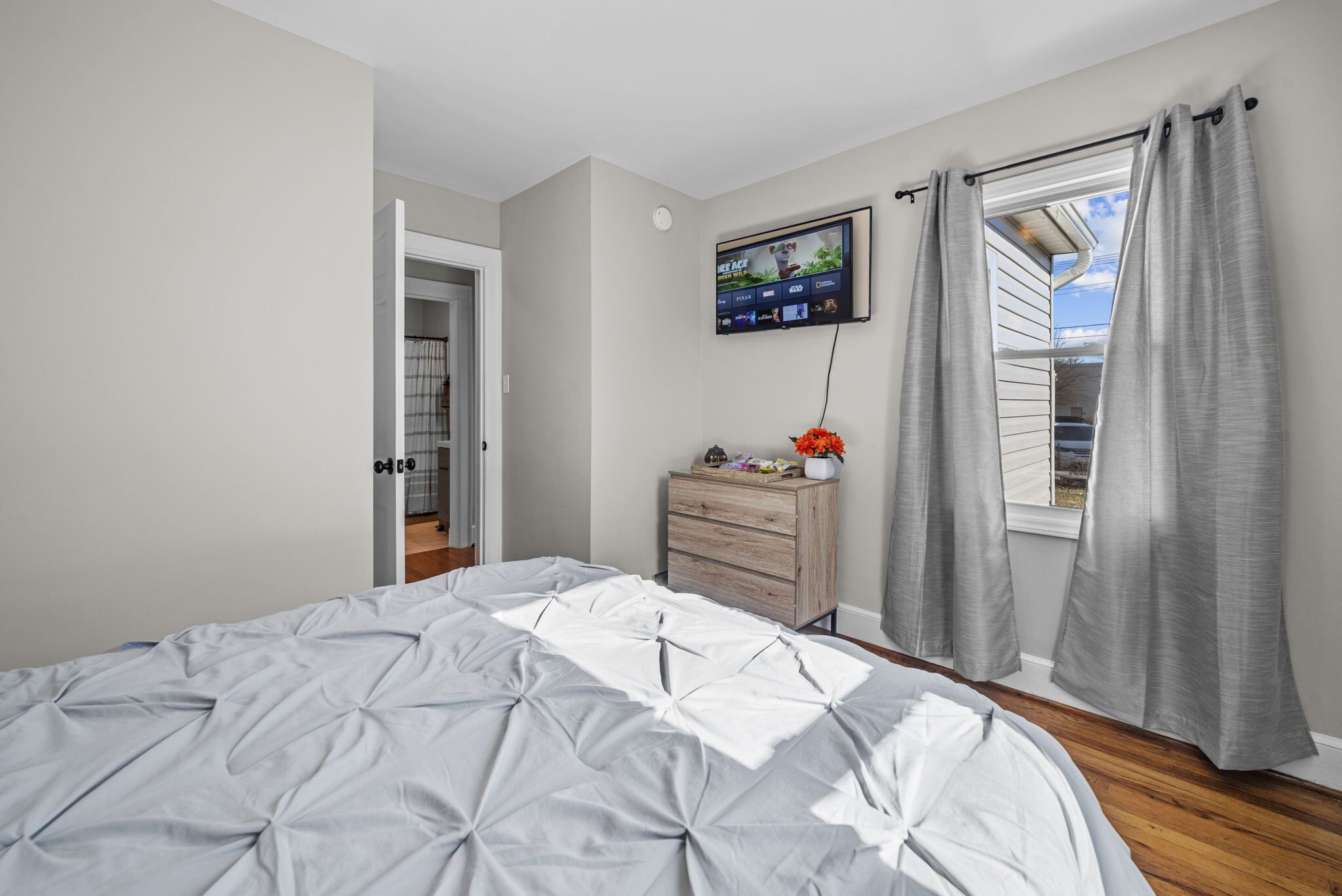 Une chambre avec un grand lit, un téléviseur fixé au mur, une commode et une fenêtre avec des rideaux.