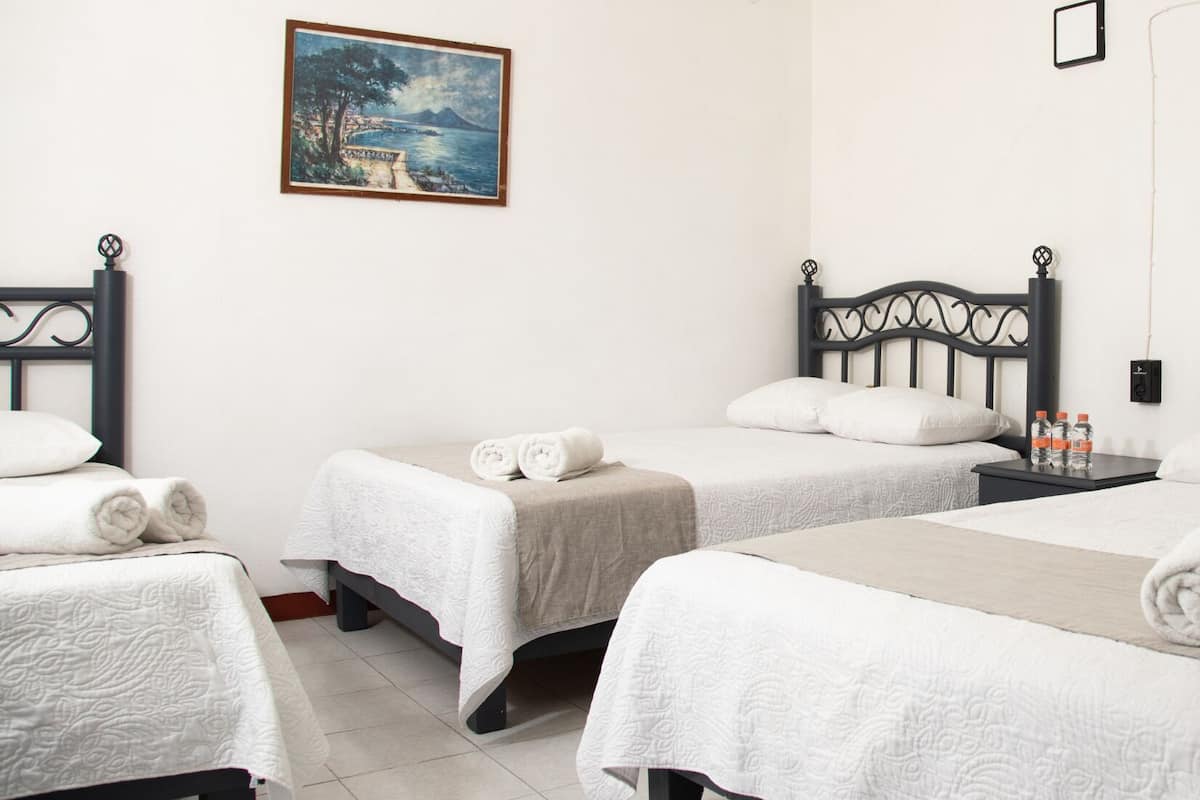 A hotel room with three beds, each with a white bedspread and rolled towels. A painting of a coastal landscape hangs on the wall.