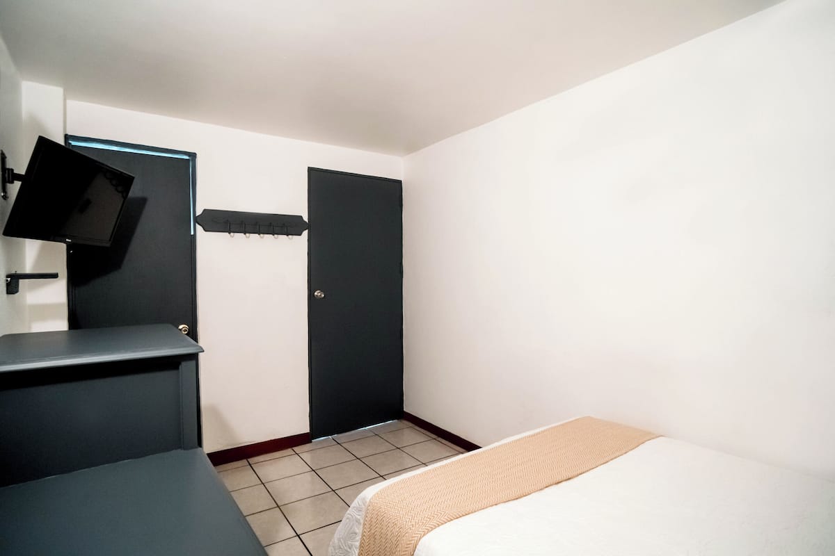 A small, clean room with a bed, a desk, a chair, a TV mounted on the wall, and a door.