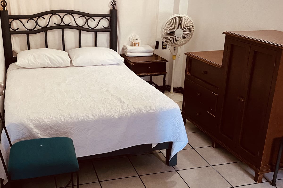 A neatly made bed with a white comforter and pillows, a bedside table with a fan, a wooden dresser, and a green chair.