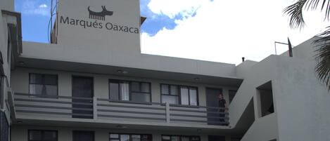Un edificio con un letrero que dice "Marqués Oaxaca", con mesas y sillas en la terraza, y un cielo despejado con nubes.
