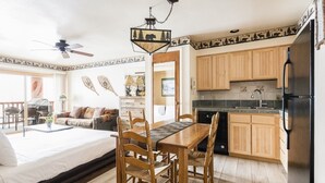 Un dormitorio con cama, una mesa de comedor con sillas, una cocina con armarios de madera y un refrigerador, y un ventilador de techo.
