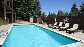 A rectangular swimming pool with clear blue water, surrounded by a concrete deck. There are four lounge chairs with cushions, a small shed, and a wooden fence with trees in the background.