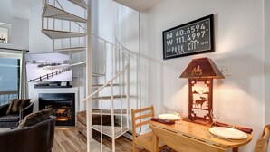Uma sala de estar moderna com escada de madeira, lareira, mesa de jantar posta para dois e televisão exibindo uma paisagem nevada.