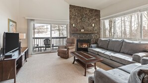A living room with a stone fireplace, a grey sofa, a recliner, a TV, and a balcony with a view of trees. - Snow Flower 130 2 Bedroom Condo by RedAwning (Park City)