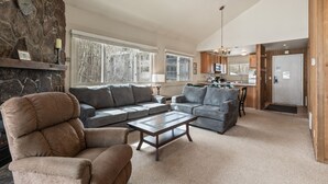 A spacious living room with a stone fireplace, large windows, and a kitchen in the background. - Snow Flower 130 2 Bedroom Condo by RedAwning (Park City)