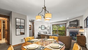 A dining area with a round table set for four, surrounded by wooden chairs. A television is mounted on the wall above the fireplace, and there is a large window providing a view of the outdoors. - Snow Flower 142 2 Bedroom Condo by RedAwning (Park City)