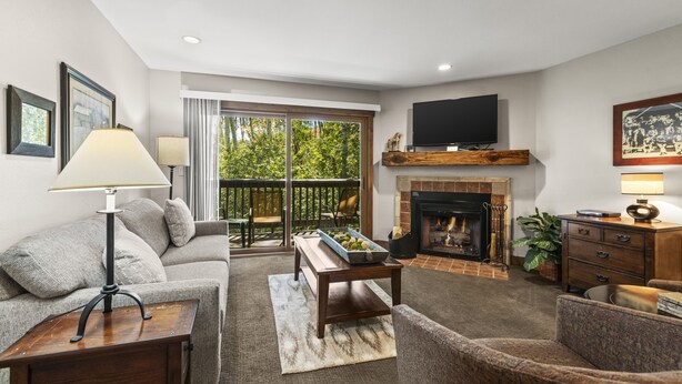 A living room with a sofa, armchair, coffee table, fireplace, television, and a balcony with a view of trees. - Snow Flower 142 2 Bedroom Condo by RedAwning (Park City)