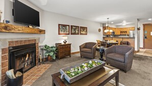 A living room with a fireplace, a flat-screen TV, and a dining area with a kitchen in the background. - Snow Flower 142 2 Bedroom Condo by RedAwning (Park City)