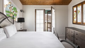 A bedroom with a wooden ceiling, a bed with white linens, a nightstand with a lamp, a dresser, and a window with a stained glass design.