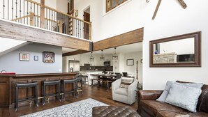 A spacious living area with a high ceiling, a wooden staircase, a kitchen with white cabinets, and a large mirror.