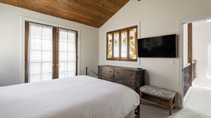 A bedroom with a large bed, a wooden headboard, a bench, a TV mounted on the wall, and a window with a stained glass design.
